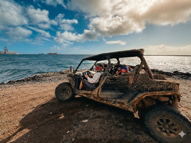 Una UTV buggy de cuatro plazas, completamente cubierta de barro, está estacionada en un camino costero rocoso y de tierra junto al océano. Se ven dos personas con gafas; el conductor está al volante y un pasajero trasero hace el signo de la paz. El fondo es un mar azul bajo un cielo dramático y nublado con luz solar brillante rompiendo en el horizonte. Se ve infraestructura costera en la distancia lejana.