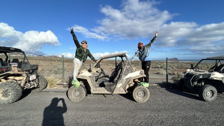 Dos personas posan de pie sobre las ruedas de un buggy todoterreno embarrado, haciendo signos de paz y victoria, en un paisaje desértico costero con vista al mar y otras buggies, bajo un cielo azul con nubes.