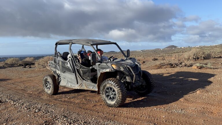 Un buggy UTV de cuatro plazas circula por un sendero desértico polvoriento y rocoso con cuatro personas con gafas protectoras en su interior. El pasajero delantero hace el signo de la paz y el fondo muestra un terreno árido y montañoso, una ciudad lejana y un cielo nublado.